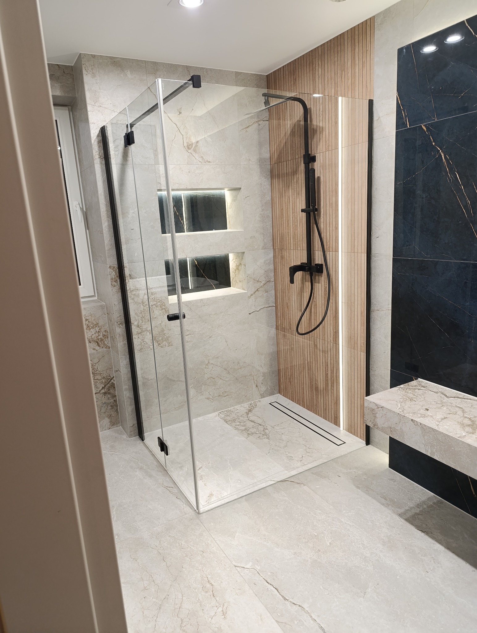 Nowoczesna kabina prysznicowa z czarnymi detalami i odpływem liniowym, wykończona płytkami imitującymi marmur i drewno. W tle wnęki z oświetleniem.