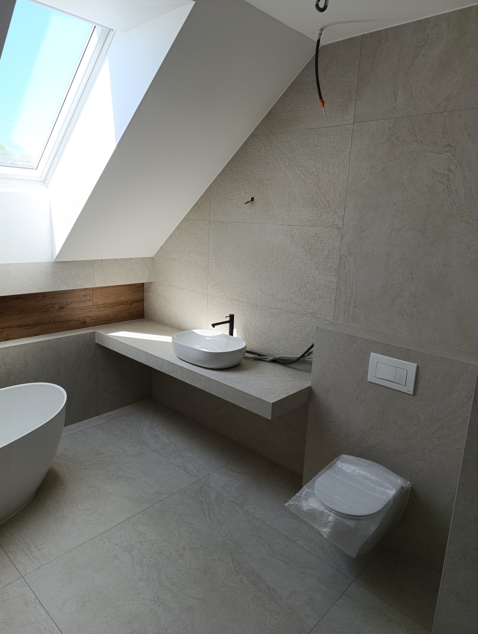 Jasna łazienka na poddaszu z oknem dachowym. Umywalka nablatowa, beżowe płytki imitujące kamień, wanna wolnostojąca. Toaleta zabezpieczona folią.