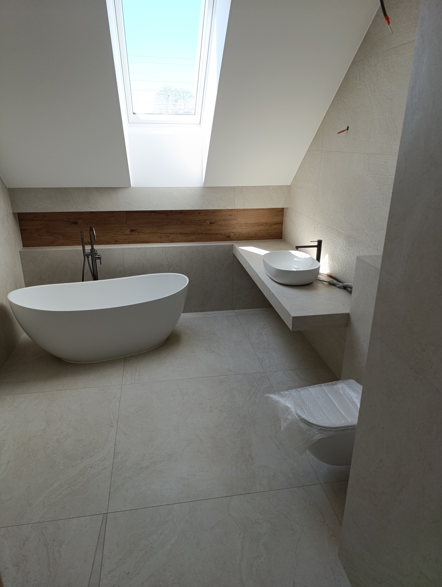 Nowoczesna łazienka na poddaszu z wolnostojącą wanną, umywalką nablatową i oknem dachowym. Beżowe płytki imitujące kamień. Umywalka z czarną armaturą.