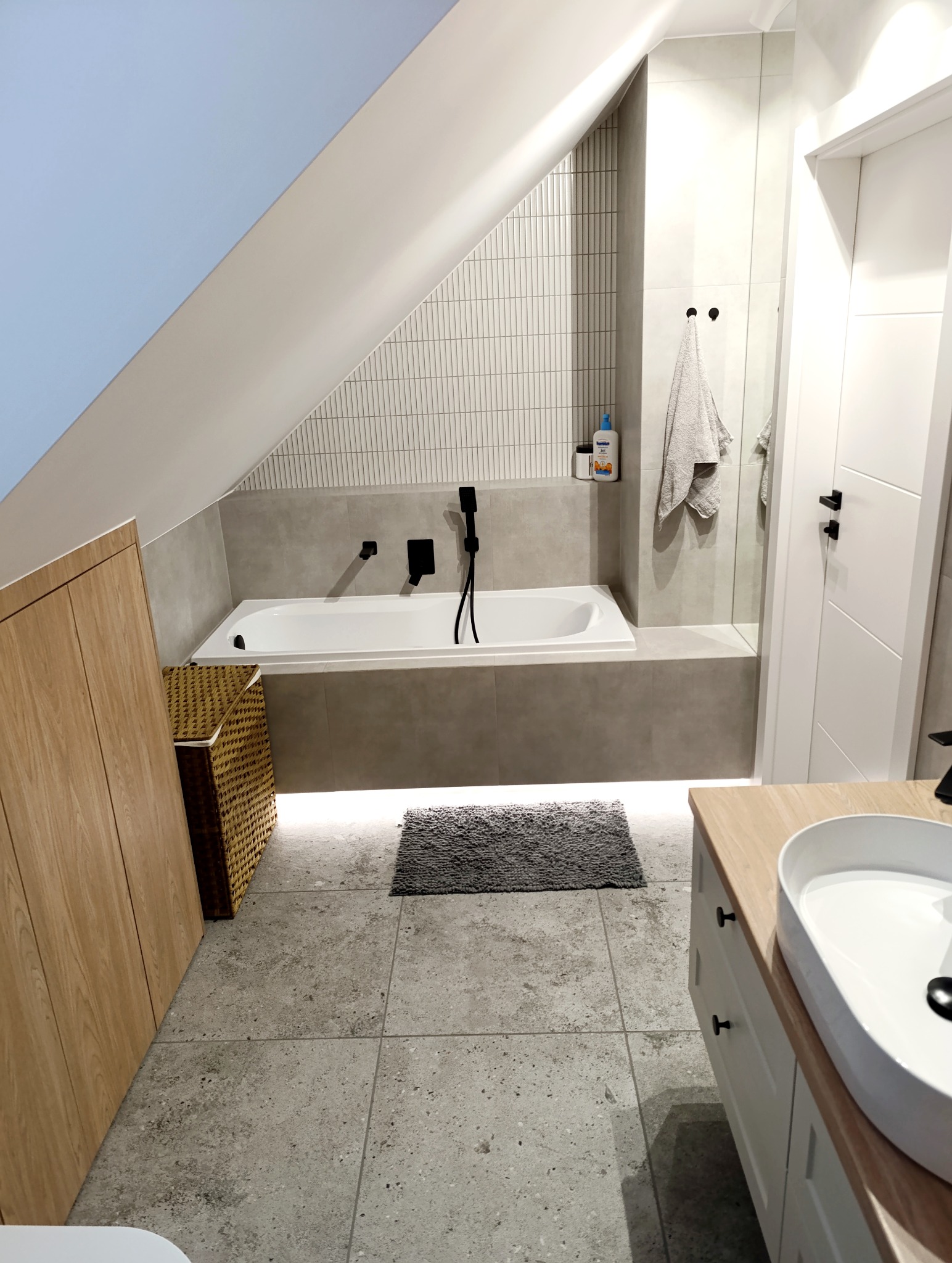 Nowoczesna łazienka na poddaszu w Kętach. Biała wanna z czarną armaturą, szare płytki i drewniane szafki tworzą spójną całość. Uwagę przykuwa oświetlenie LED pod wanną.