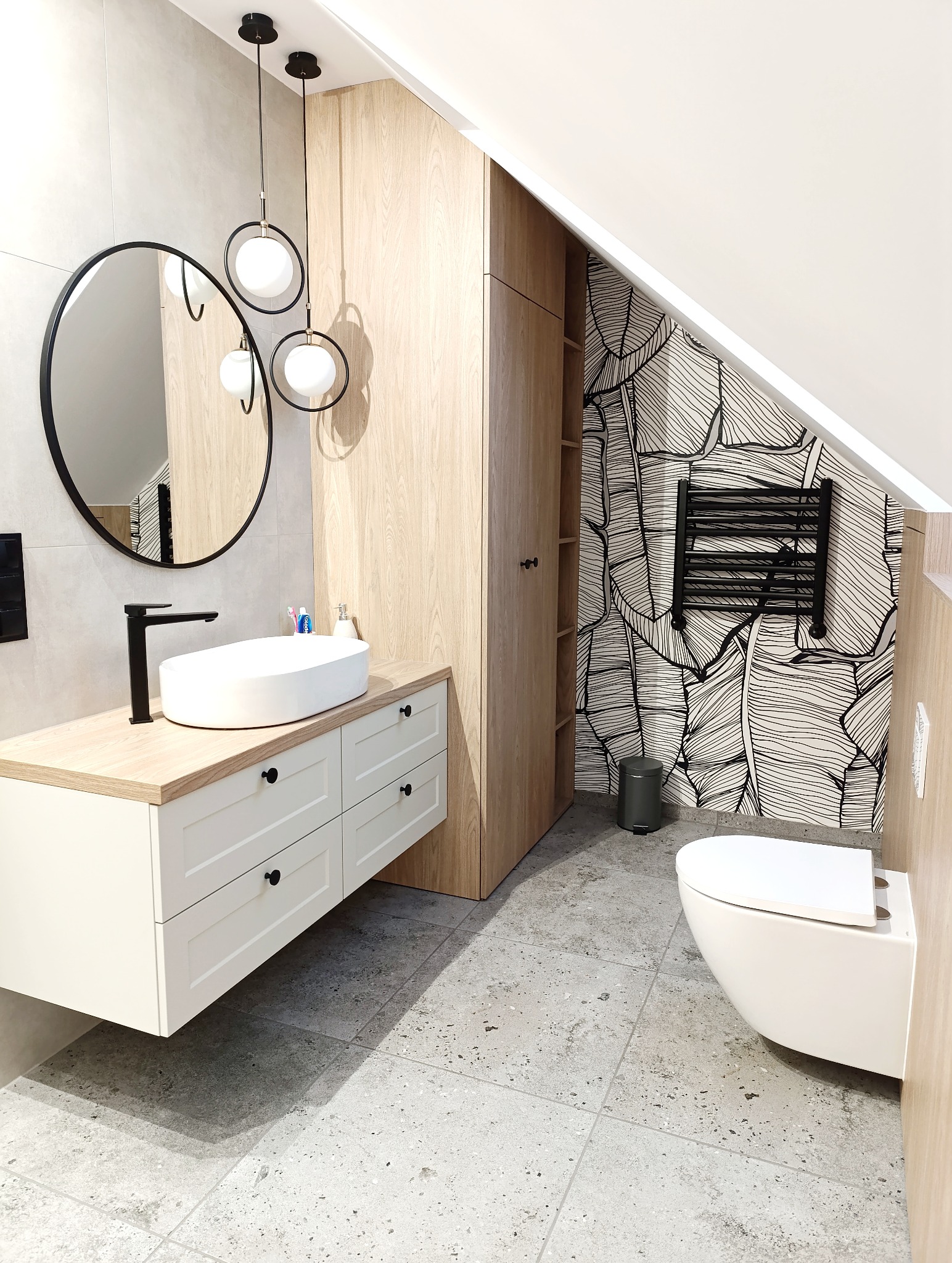 Nowoczesna łazienka na poddaszu z umywalką nablatową, okrągłym lustrem, szafką z jasnego drewna i wzorzystą tapetą. Grzejnik drabinkowy i wisząca toaleta.