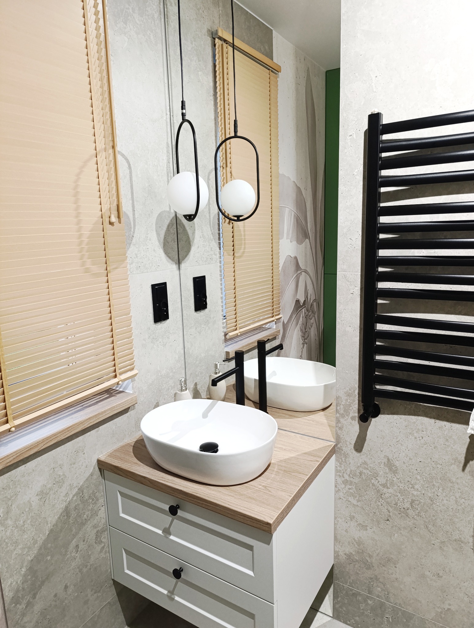 Nowoczesna łazienka z owalną umywalką nablatową, czarną armaturą i grzejnikiem drabinkowym. Jasne rolety i designerskie lampy kuliste dopełniają aranżację.