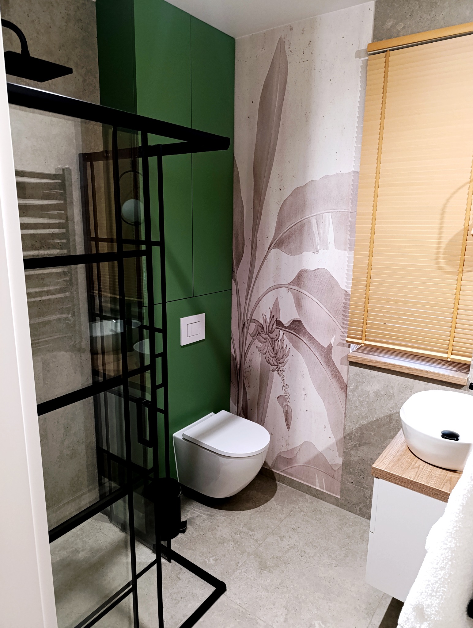 Nowoczesna łazienka z kabiną typu walk-in, zielonymi szafkami, toaletą podwieszaną i umywalką nablatową. Na ścianie tapeta z motywem roślinnym i drewniane żaluzje.