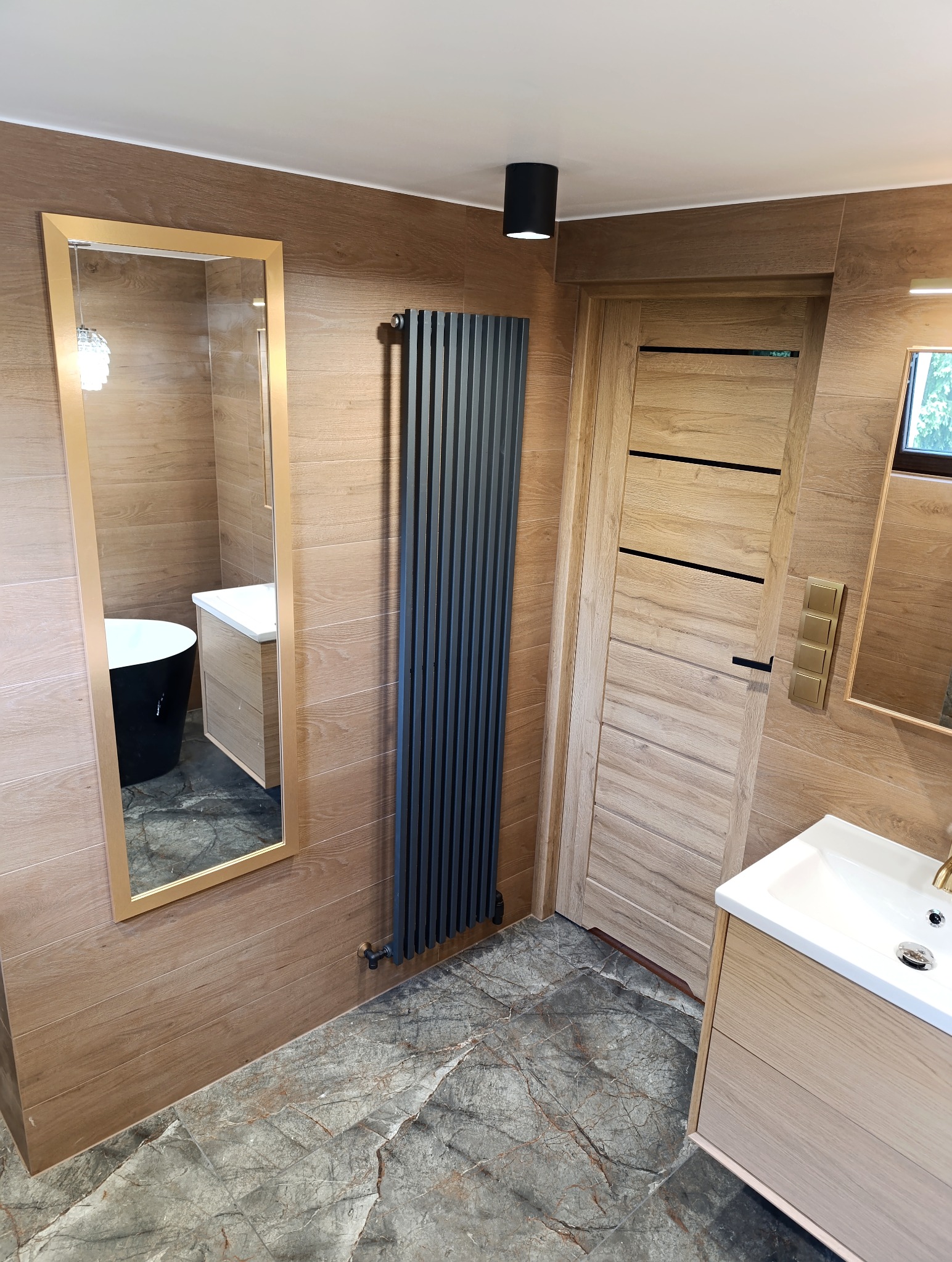 Nowoczesna łazienka z drewnopodobnymi płytkami, szarym grzejnikiem panelowym i marmurową posadzką. W lustrze odbija się czarna wanna wolnostojąca.
