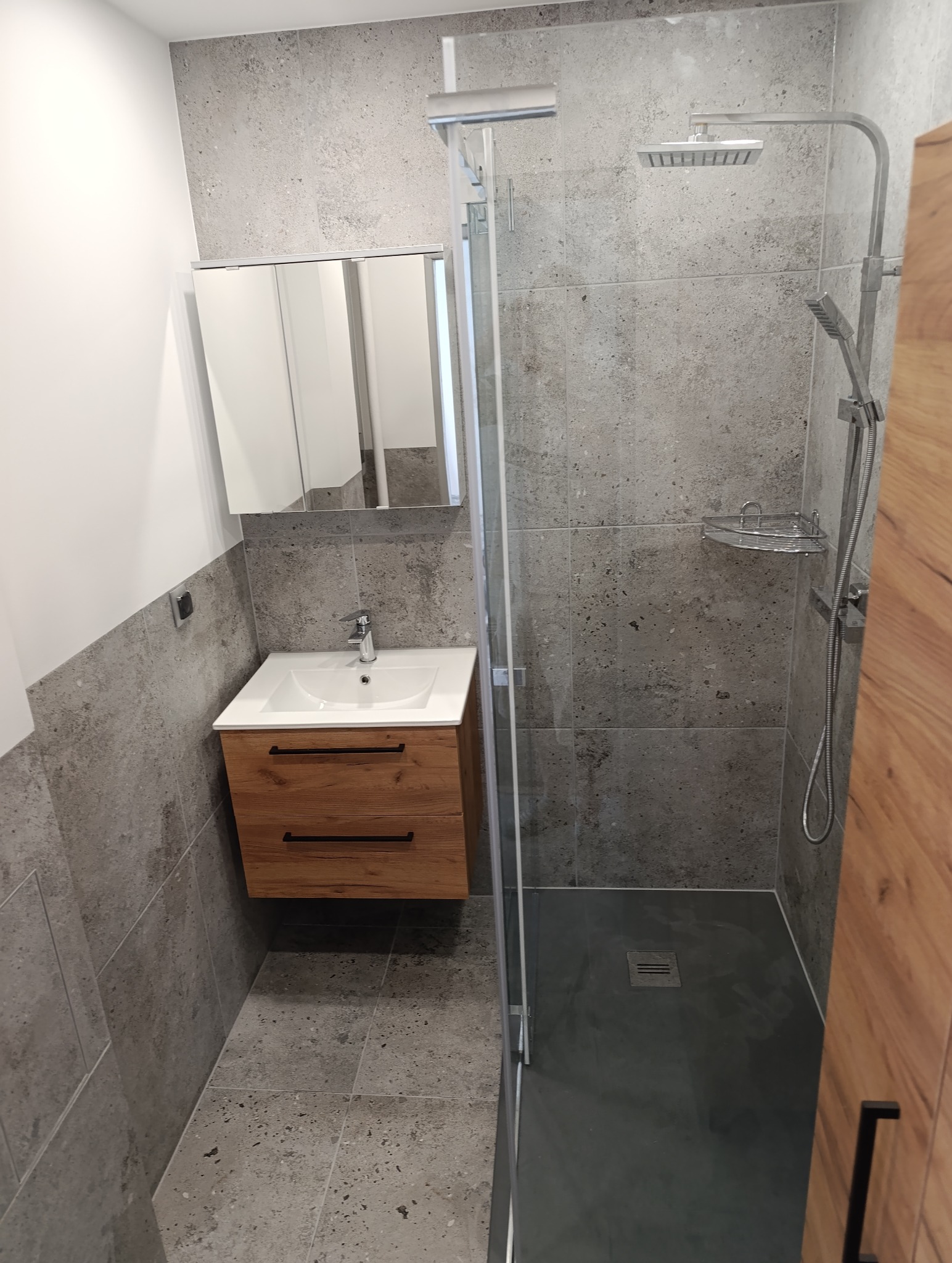 Nowoczesna łazienka po remoncie: szare płytki imitujące beton, drewniana szafka pod umywalką, kabina prysznicowa ze szklaną ścianką i deszczownicą.