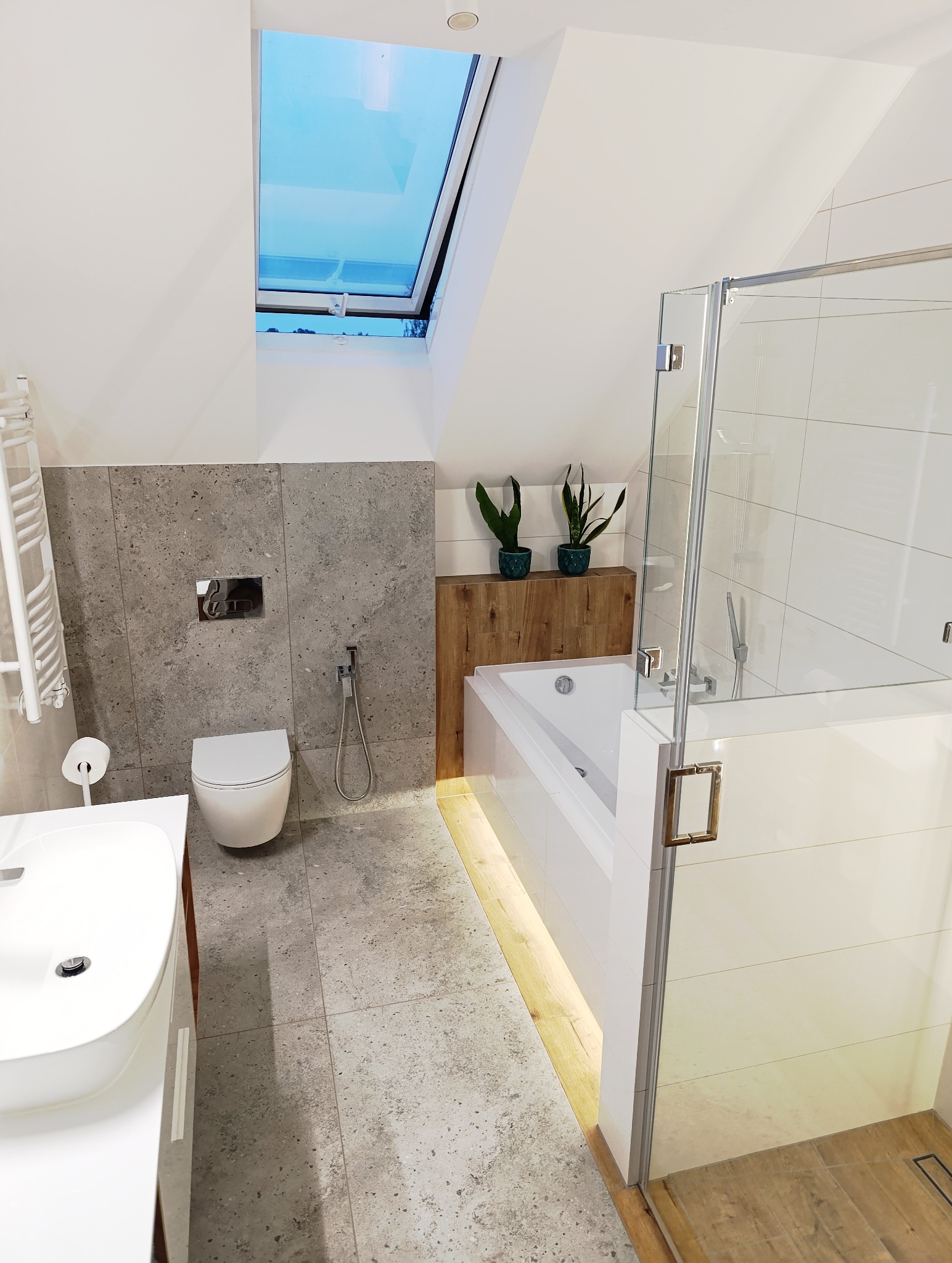Jasna łazienka na poddaszu z oknem dachowym. Szare płytki, biała armatura, kabina prysznicowa ze złotymi uchwytami. Dwie rośliny w doniczkach nad wanną.