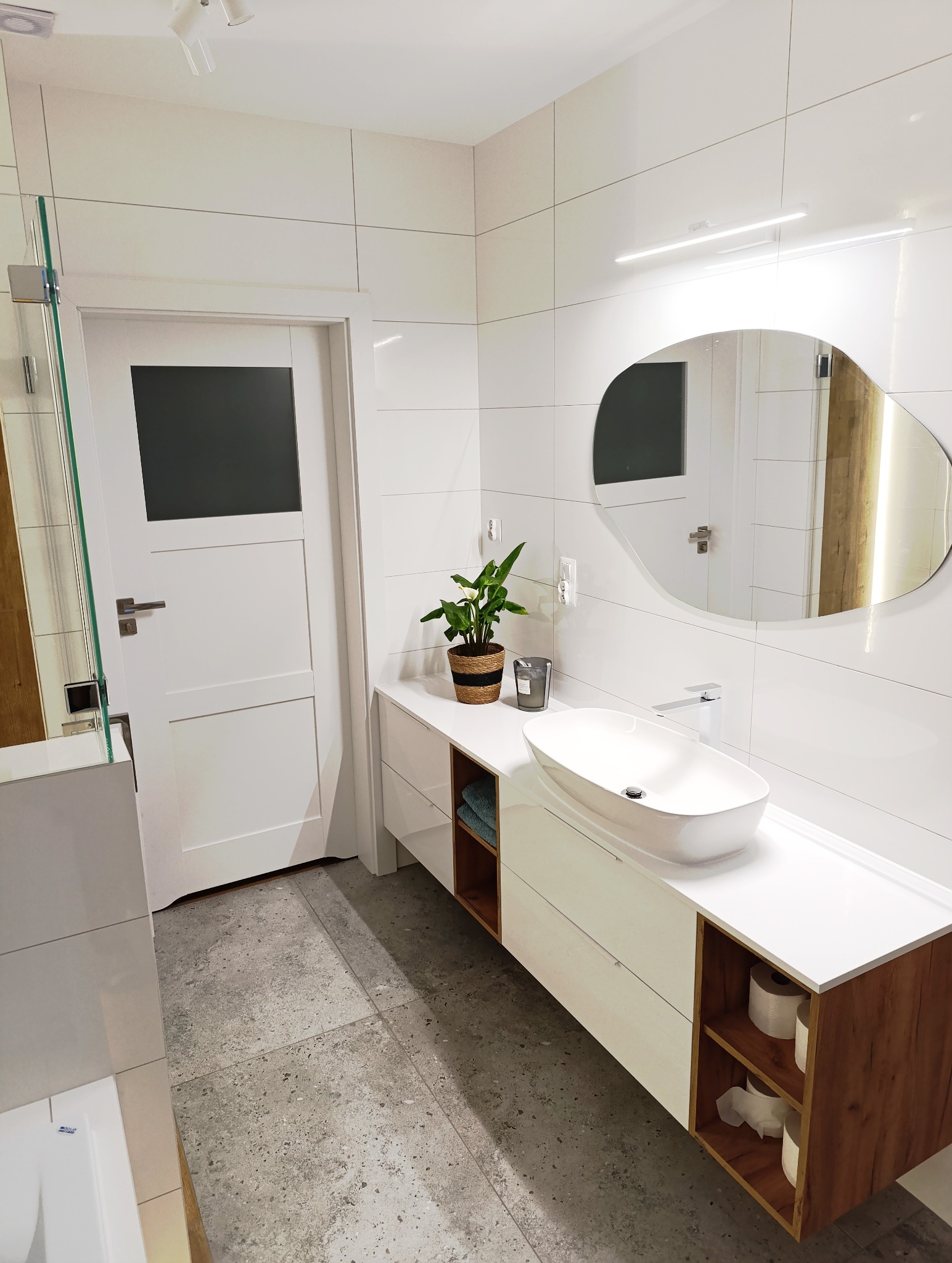 Nowoczesna łazienka z białą armaturą, okrągłym lustrem z podświetleniem i szarą podłogą. Uwagę przykuwa roślina w ozdobnej doniczce na blacie umywalki.