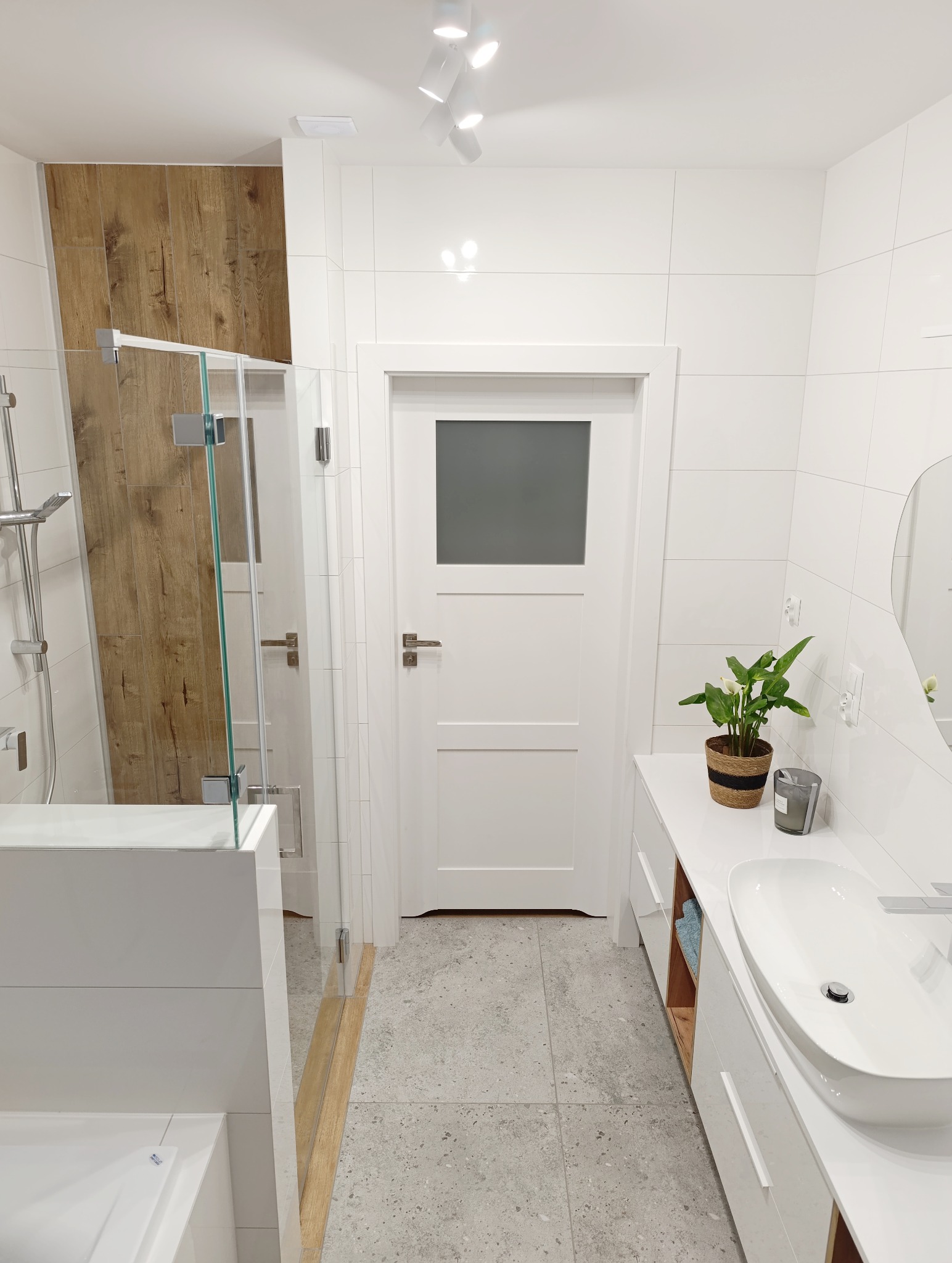 Jasna łazienka z kabiną prysznicową, drewnianym akcentem na ścianie, białą umywalką nablatową i rośliną doniczkową. Minimalistyczny design, nowoczesne oświetlenie.