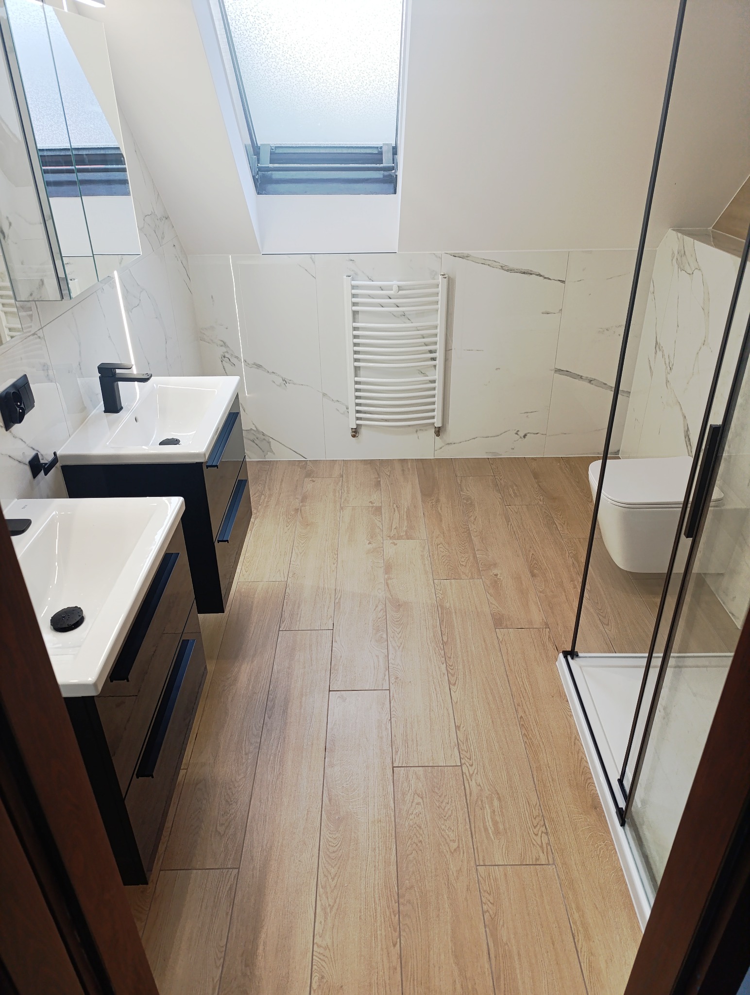 Nowoczesna łazienka na poddaszu z drewnopodobną podłogą, biało-marmurowymi płytkami ściennymi, kabiną prysznicową z czarnymi ramami i podwójną umywalką z czarną armaturą.