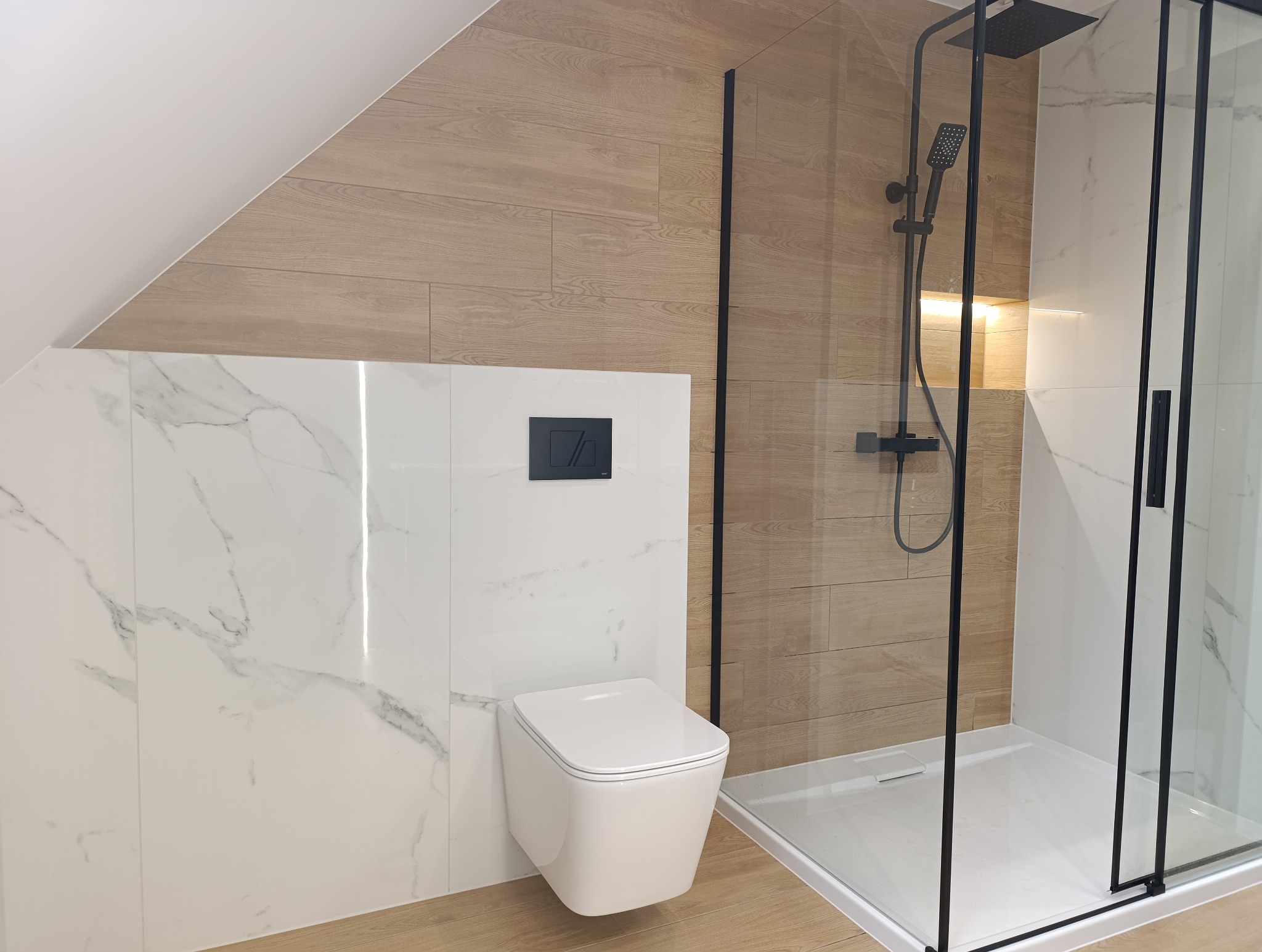 Nowoczesna łazienka na poddaszu z kabiną prysznicową typu walk-in, białymi płytkami imitującymi marmur i drewnopodobnymi płytkami na skośnej ścianie.