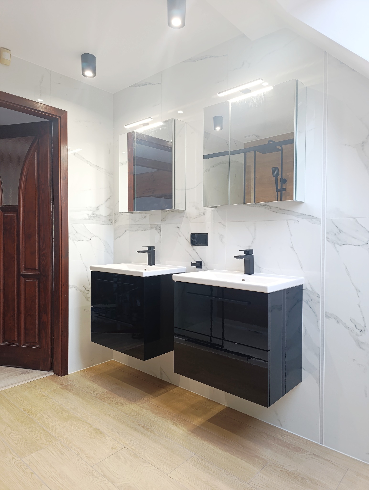 Nowoczesna łazienka z dwoma czarnymi szafkami pod umywalki, lustrami z oświetleniem i marmurowymi płytkami. Widoczne drewniane drzwi i podłoga imitująca drewno.