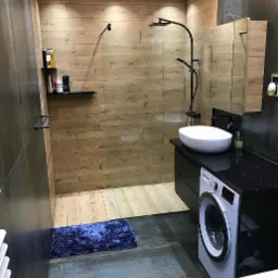 Nowoczesna łazienka z kabiną prysznicową typu walk-in, wykończona drewnopodobnymi płytkami, z czarną armaturą, umywalką nablatową, pralką i ciemnoniebieskim dywanikiem.