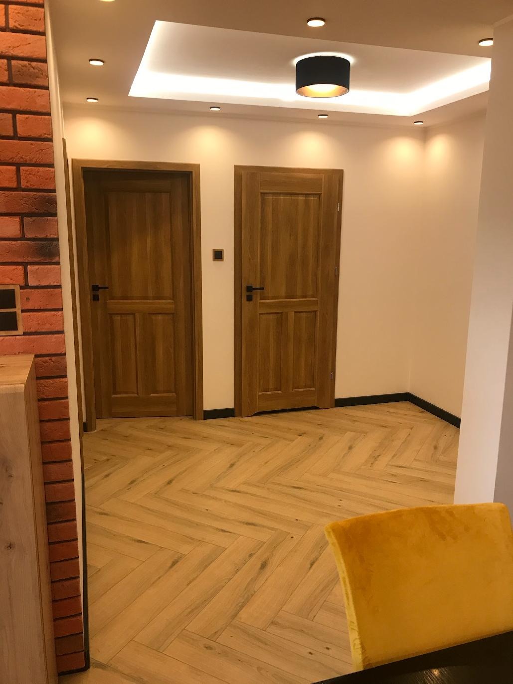 Wykończone wnętrze: Dwie drewniane drzwi wewnętrzne z czarnymi klamkami, ułożona w jodełkę jasna podłoga z paneli, ceglana ściana w tle, czarna lampa sufitowa.