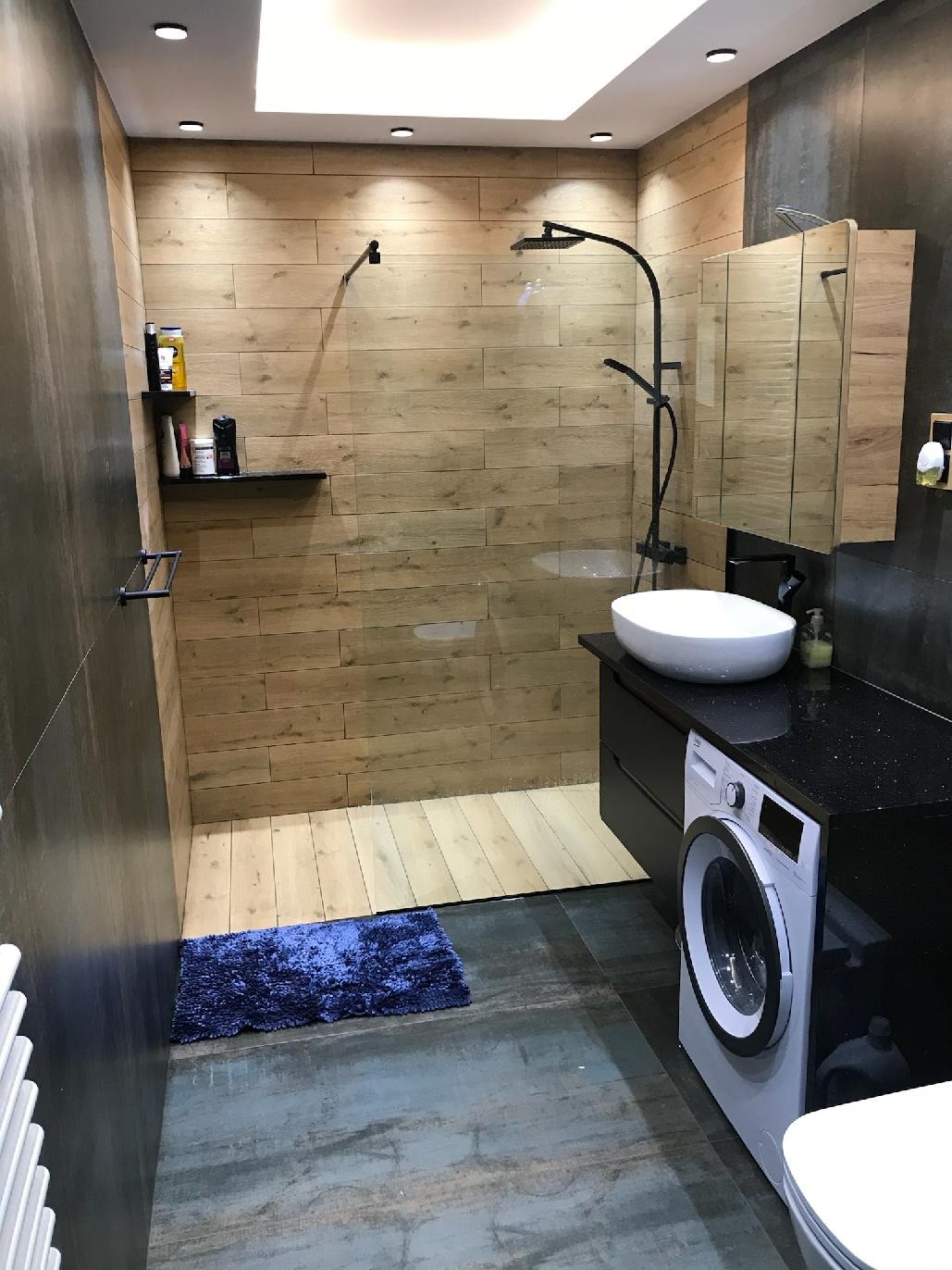 Nowoczesna łazienka z kabiną prysznicową typu walk-in, wykończona drewnopodobnymi płytkami, z czarną armaturą, umywalką nablatową, pralką i ciemnoniebieskim dywanikiem.
