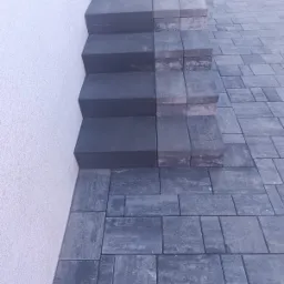 Ukończone schody z kostki brukowej, w odcieniach szarości, prowadzące wzdłuż białej ściany budynku. Widoczny fragment chodnika z tej samej kostki.