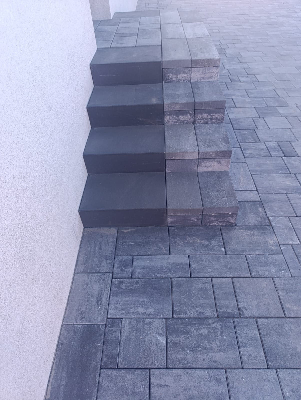 Ukończone schody z kostki brukowej, w odcieniach szarości, prowadzące wzdłuż białej ściany budynku. Widoczny fragment chodnika z tej samej kostki.