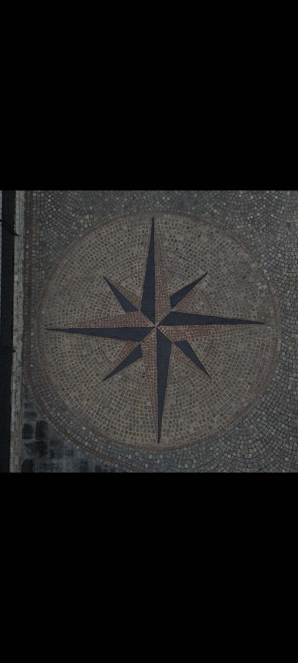 Mozaika brukowa z wzorem kompasu, widok z góry. Ciemne, gładkie kostki tworzą gwiazdę na tle szaro-brązowych, drobnych kamieni. Detal wykończenia nawierzchni.