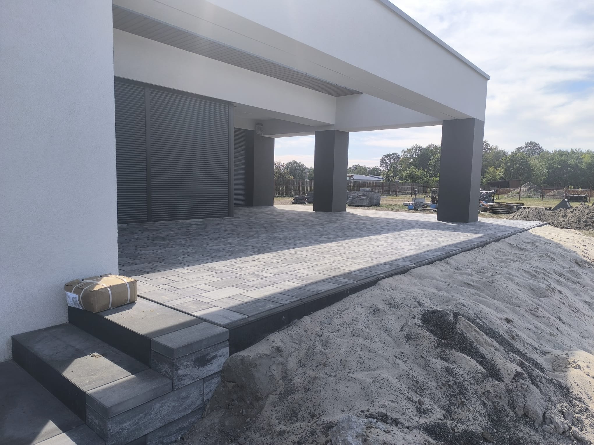 Nowoczesny taras z szarej kostki brukowej ułożonej w geometryczny wzór, widoczne schody wejściowe i przygotowany teren pod dalsze prace.