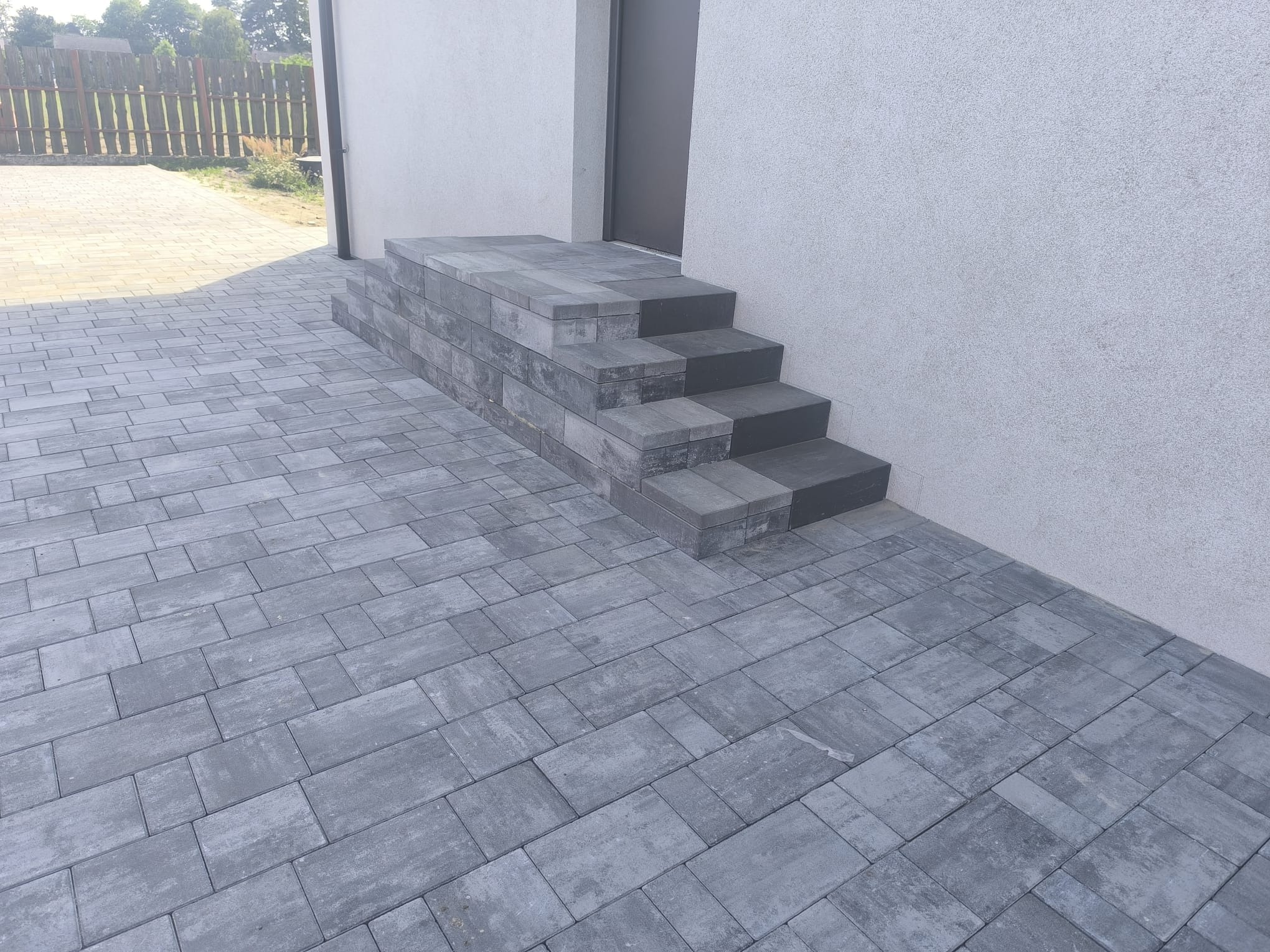 Szare schody z kostki brukowej prowadzące do drzwi budynku, obok ułożona kostka brukowa. Widok na fragment ogrodzenia w tle. Minimalistyczny styl.