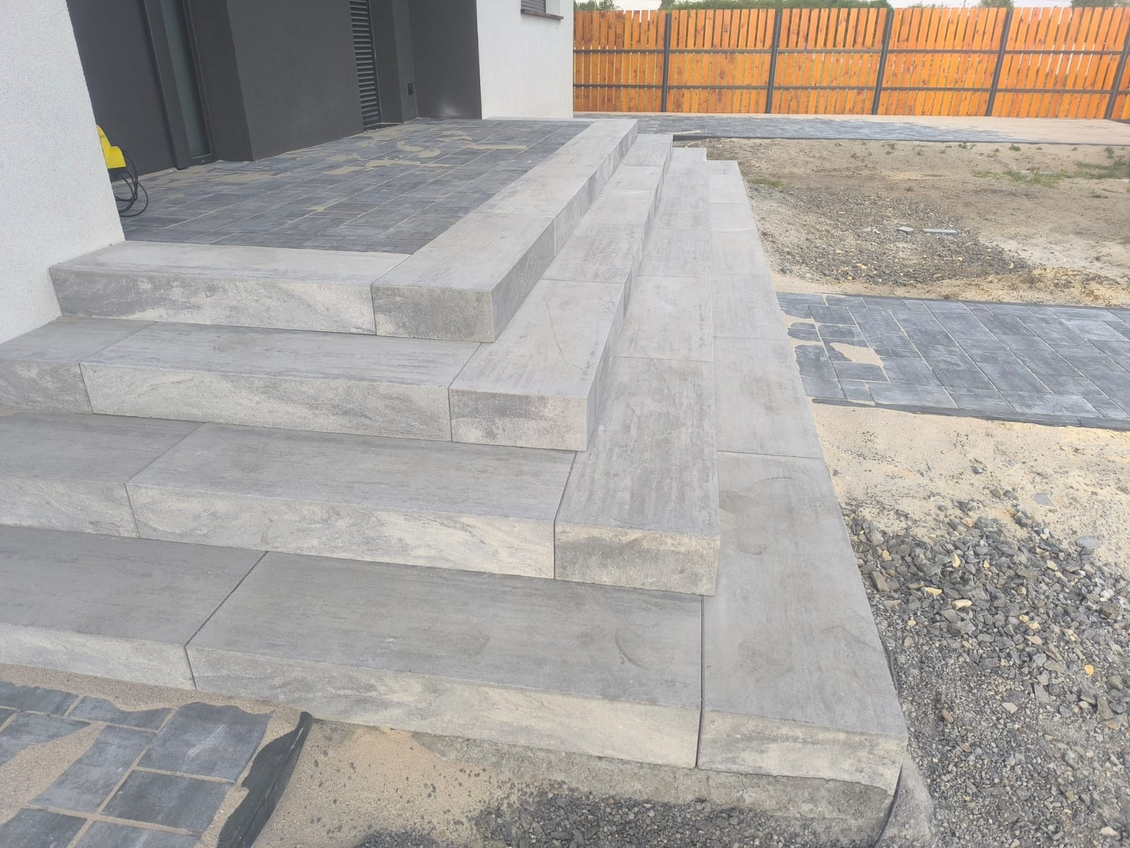 Schody zewnętrzne z szarej kostki brukowej, prowadzące do domu. Widoczne także fragmenty chodnika z kostki i drewnianego ogrodzenia w tle.