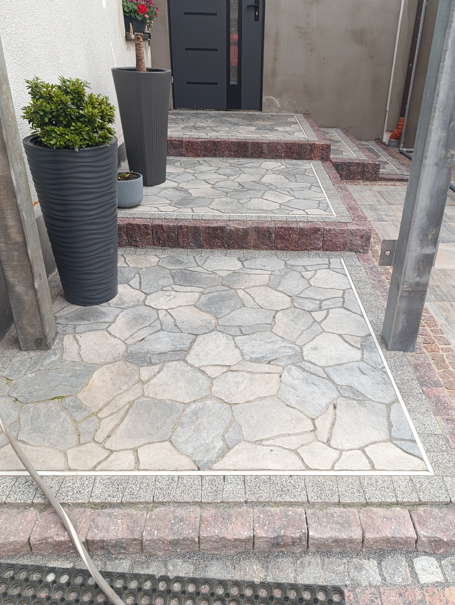 Ułożona kostka brukowa o nieregularnym kształcie, obrzeża z czerwonej kostki, donice z roślinami przy wejściu do domu. Widok na schody wejściowe.