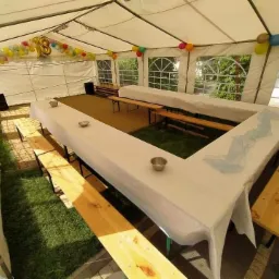 Wnętrze białego namiotu imprezowego z drewnianymi ławami i stołami w kształcie litery U, nakrytymi białymi obrusami, dekorowane kolorowymi balonami i cyfrą 18, widok z góry na trawnik i kostkę...