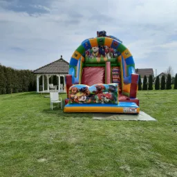 Kolorowy dmuchany zamek ze zjeżdżalnią z motywem bajki Psi Patrol, ustawiony na trawniku obok białej altany ogrodowej.