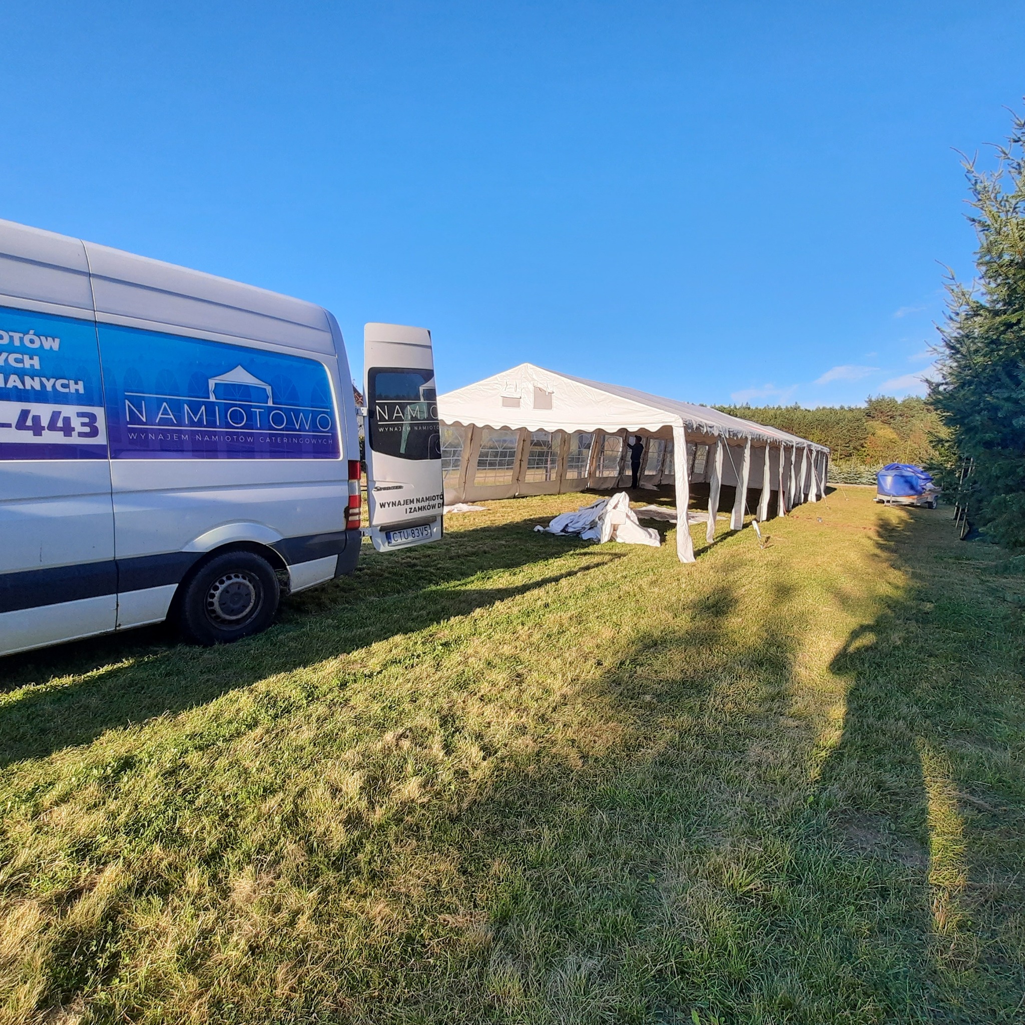 Rozstawiony biały namiot na trawie, częściowo widoczny samochód dostawczy z logo firmy oferującej wynajem namiotów i catering, błękitne niebo w tle.