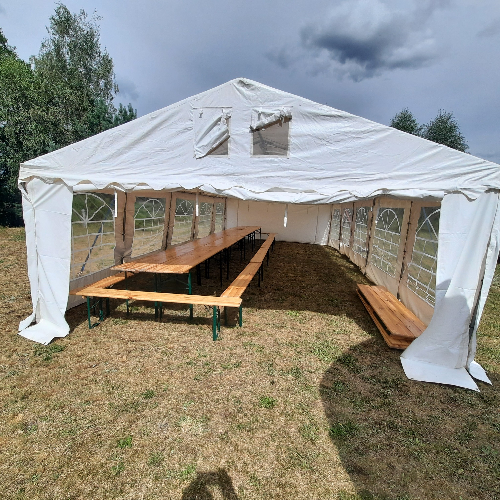 Biały namiot imprezowy z drewnianymi stołami i ławkami w środku, rozłożony na trawie, widok z zewnątrz przy pochmurnym niebie.