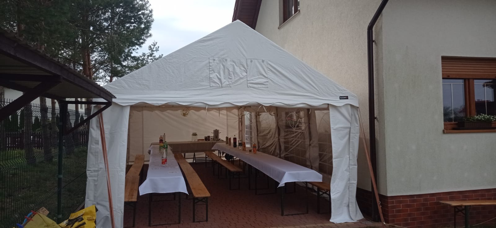Biały namiot plenerowy rozstawiony przy budynku z oknem, wewnątrz dwa długie stoły z białymi obrusami i ławkami, przygotowane na imprezę.