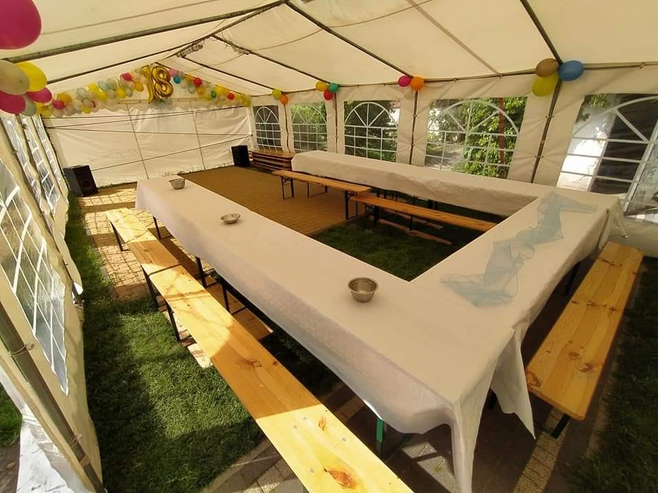 Wnętrze białego namiotu imprezowego z drewnianymi ławami i stołami w kształcie litery U, nakrytymi białymi obrusami, dekorowane kolorowymi balonami i cyfrą 18, widok z góry na trawnik i kostkę...