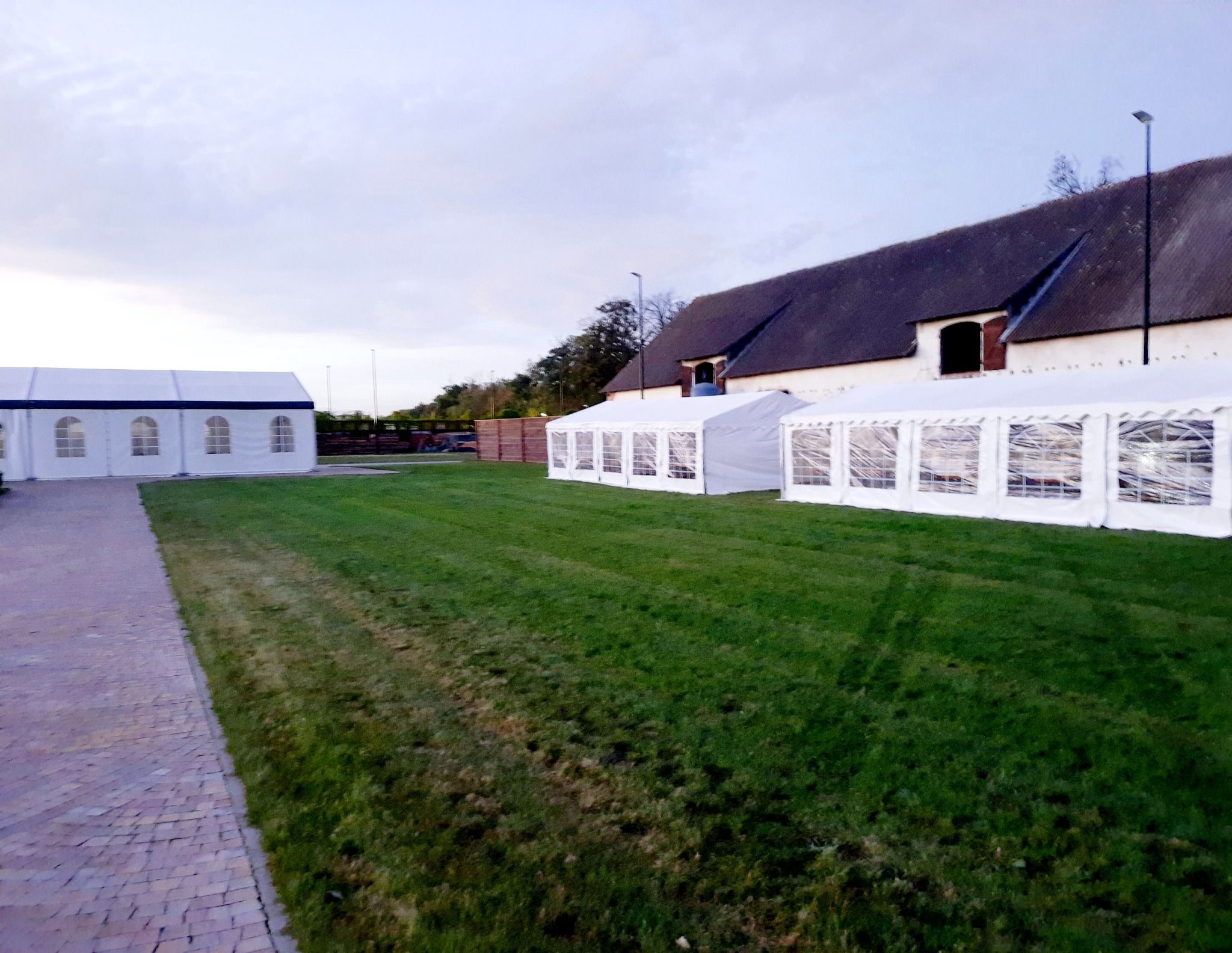 Rozstawione białe namioty z oknami na trawniku przed budynkiem z ciemnym dachem, widok z boku, pochmurne niebo.