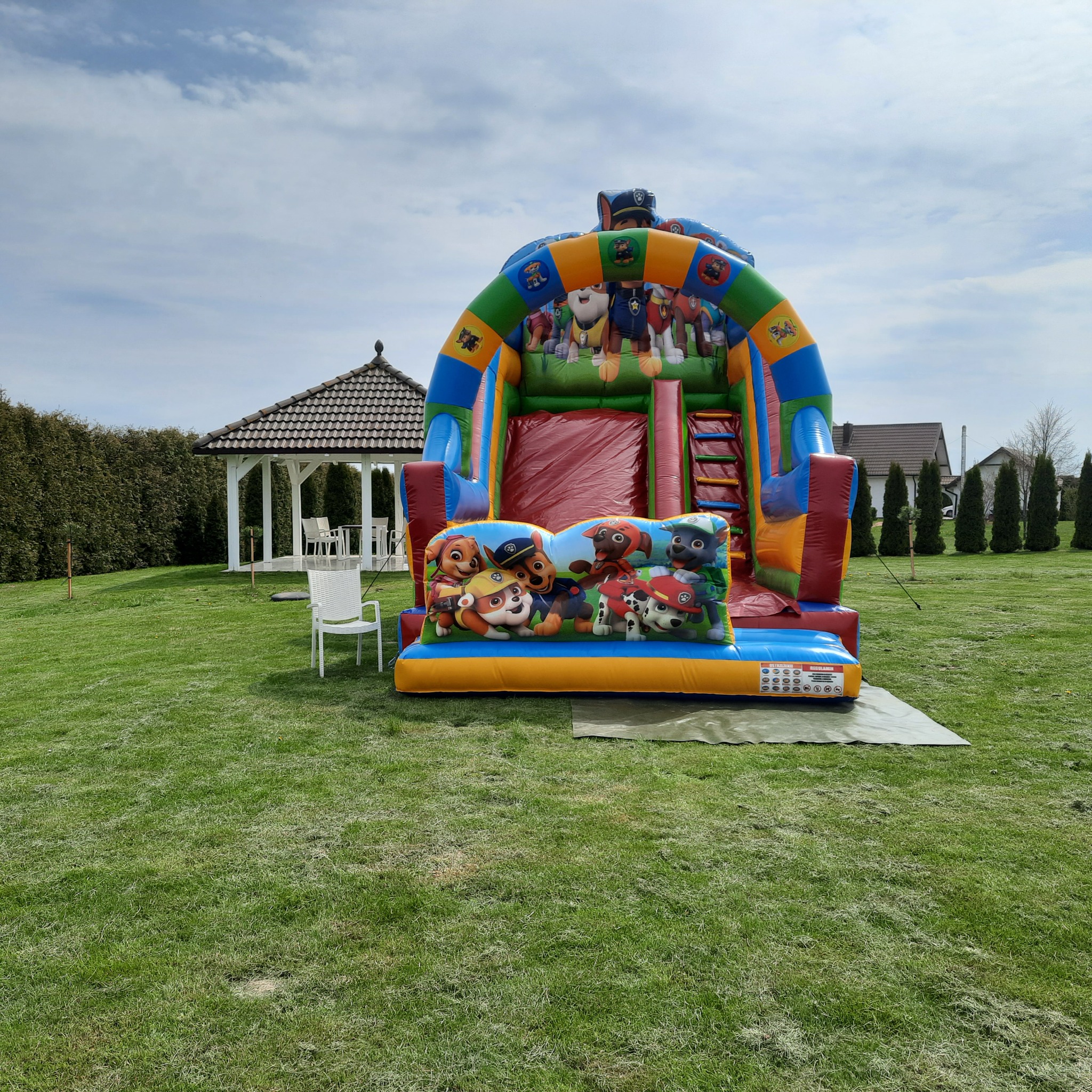 Kolorowy dmuchany zamek ze zjeżdżalnią z motywem bajki Psi Patrol, ustawiony na trawniku obok białej altany ogrodowej.