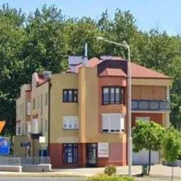 Fasada budynku prywatnej kliniki z balkonem i widocznym szyldem informacyjnym, otoczona zielenią i latarniami ulicznymi.