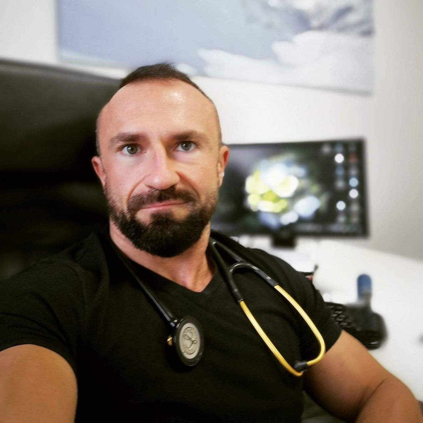 Portret lekarza ze stetoskopem w Pabianicach, z lekkim uśmiechem i skupionym spojrzeniem, siedzącego przed komputerem z rozmytym tłem biurowym.