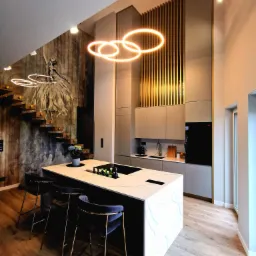 Nowoczesna kuchnia z wyspą, blatem z białego marmuru, złotymi krzesłami barowymi i schodami z motywem baletnicy na ścianie.