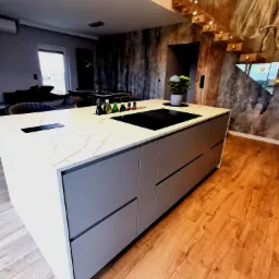 Wyspa kuchenna z blatem imitującym marmur, z wbudowaną płytą indukcyjną i gniazdkami, na drewnianej podłodze, w tle schody z drewnianymi stopniami i szklanymi balustradami.