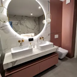 Nowoczesna łazienka z dwoma białymi umywalkami nablatowymi, marmurowym blatem, szafką w odcieniach różu, okrągłym lustrem z podświetleniem LED i złotymi bateriami oraz wiszącymi lampami.