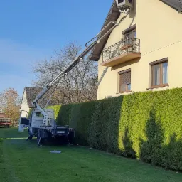 Żółta elewacja domu jednorodzinnego z ciemnym dachem, do której dostawiono podnośnik koszowy z pracownikiem w koszu na wysokości dachu.
