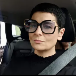 Kobieta z krótkimi włosami w okularach z grubymi oprawkami i złotymi detalami, siedząca w samochodzie z zapiętym pasem bezpieczeństwa.