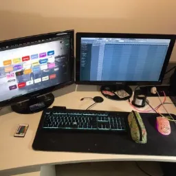 Stanowisko pracy z dwoma monitorami, klawiaturą podświetlaną na niebiesko, myszką i podkładką, widoczne naklejki na obudowie komputera.