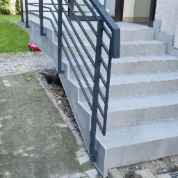 Zewnętrzne schody wejściowe wyłożone szarą płytką ceramiczną, z czarną, metalową balustradą, prowadzące do drzwi wejściowych, obok kostka brukowa i rząd tuj.