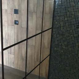 Usługi Budowlane Marek Polak - Kabina prysznicowa z czarnymi profilami, drewnopodobne płytki na ścianie z włącznikami światła i mozaika na podłodze oraz ścianie prysznica.