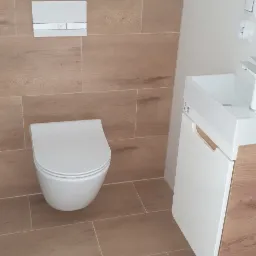 Nowoczesna łazienka z podwieszaną toaletą, umywalką z szafką z drewnianymi akcentami i płytkami imitującymi drewno na podłodze i ścianie.