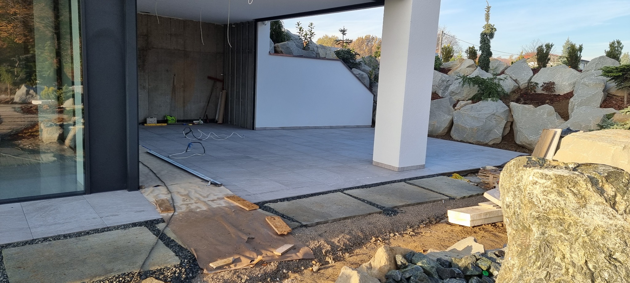 Układanie szarych płytek na tarasie w nowoczesnym domu z ogrodem skalnym w tle, widoczne narzędzia i materiały budowlane.