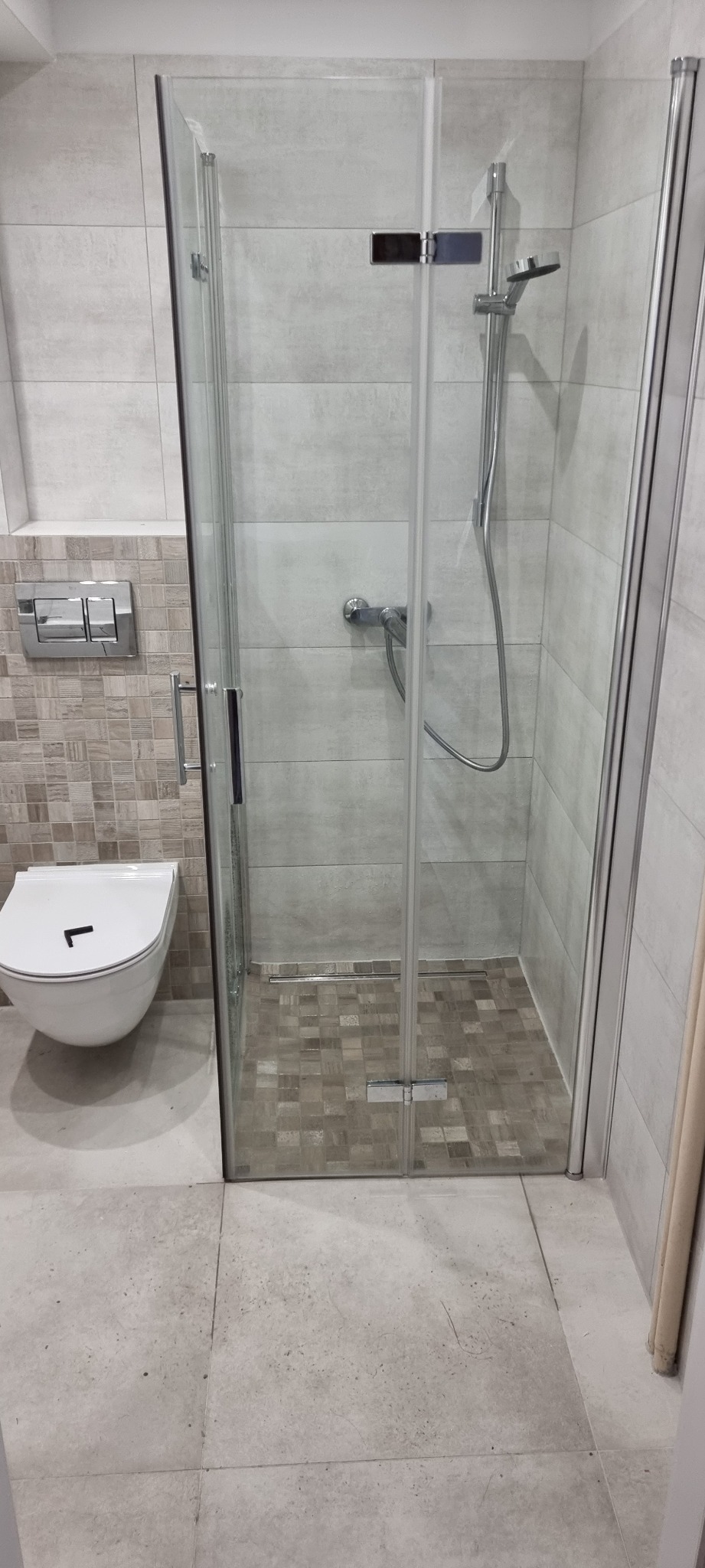 Nowoczesna kabina prysznicowa z chromowanymi elementami, jasne płytki imitujące kamień na ścianach i mozaika na podłodze wewnątrz kabiny, wisząca toaleta z chromowanym przyciskiem spłukującym obok.