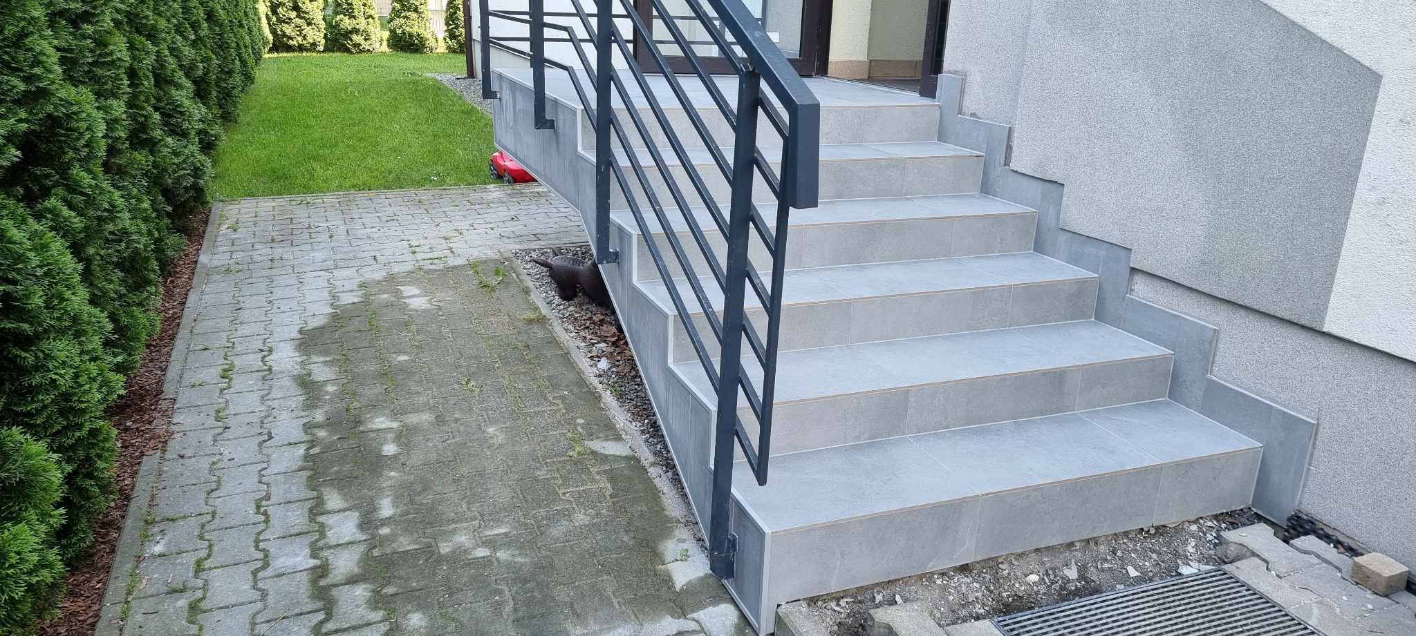 Zewnętrzne schody wejściowe wyłożone szarą płytką ceramiczną, z czarną, metalową balustradą, prowadzące do drzwi wejściowych, obok kostka brukowa i rząd tuj.