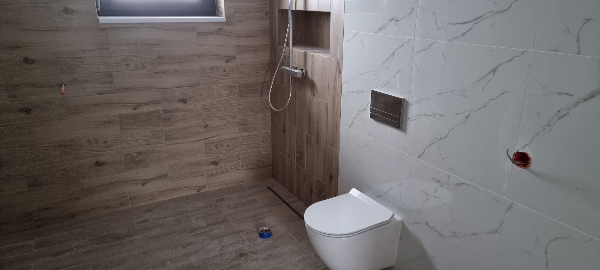Nowoczesna łazienka w trakcie wykańczania: biała toaleta podtynkowa, marmurowe płytki ścienne, drewnopodobne płytki podłogowe i ścienne w strefie prysznica, widoczne puszki elektryczne i taśma...