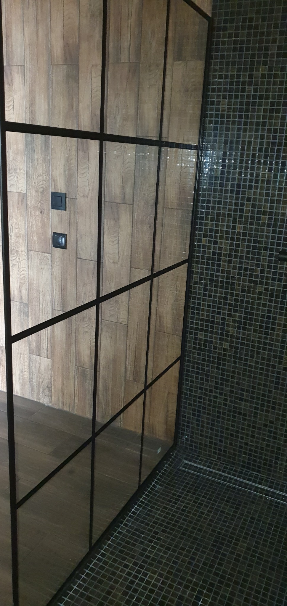 Kabina prysznicowa z czarnymi profilami, drewnopodobne płytki na ścianie z włącznikami światła i mozaika na podłodze oraz ścianie prysznica.