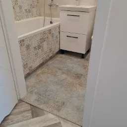 Ukończona łazienka z wanną obudowaną mozaikowymi płytkami, białą szafką z umywalką i armaturą, oraz podłogą wyłożoną wzorzystymi płytkami przechodzącymi w panele ułożone w jodełkę przy progu.